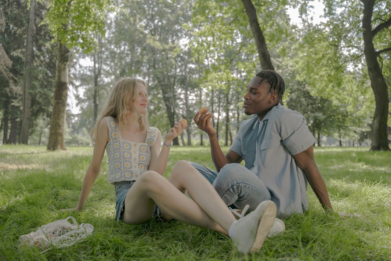 young couple sitting crossed legs on grass in park and holding fruits in hands